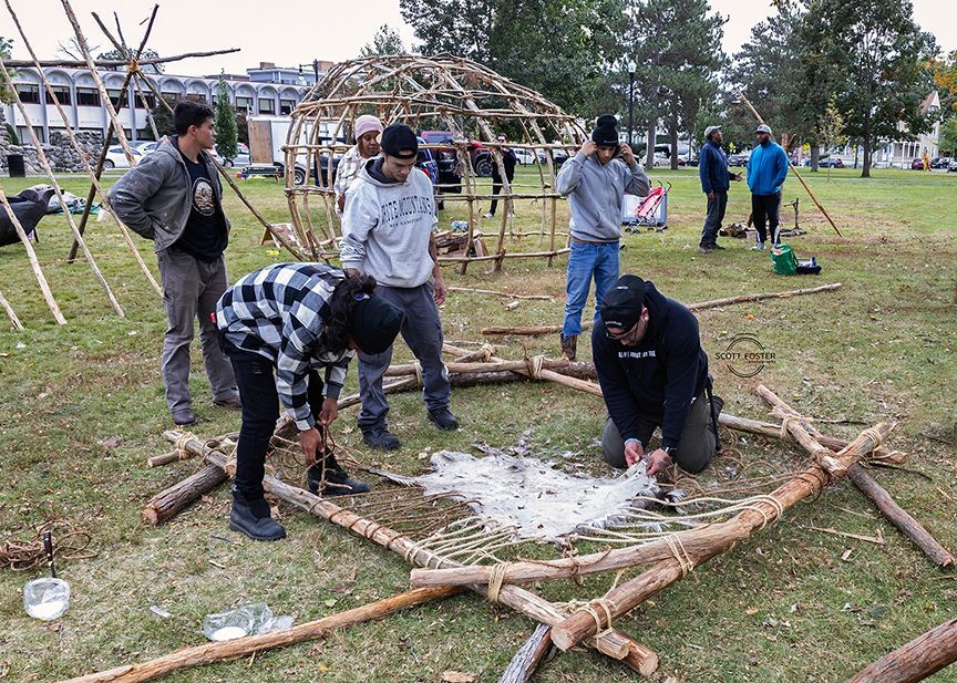 People building a wooden structure outdoors on grass. One person weaves material into the base. Others work on the frame.