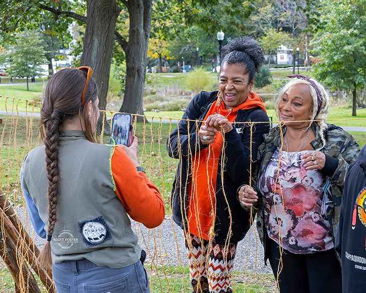 Three people outside, two women holding string art as another person photographs them.