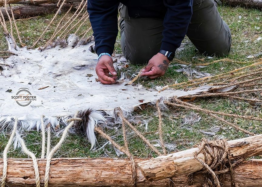 Person weaving ropes through animal hide stretched over logs outdoors.