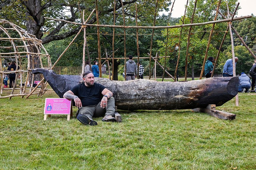 Man sits by large carved wooden whale sculpture on grass, with wooden structures in the background.