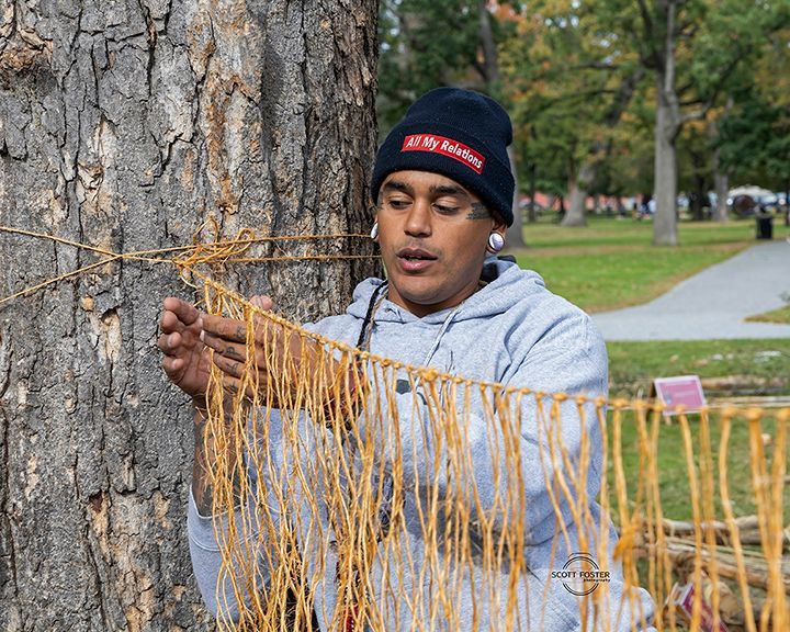 Man in beanie working with woven strands near a tree in a park.