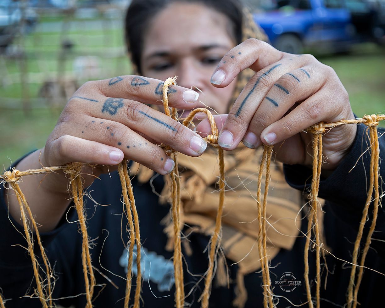 Person tying natural fibers to construct a woven structure; tattoos visible on hands.