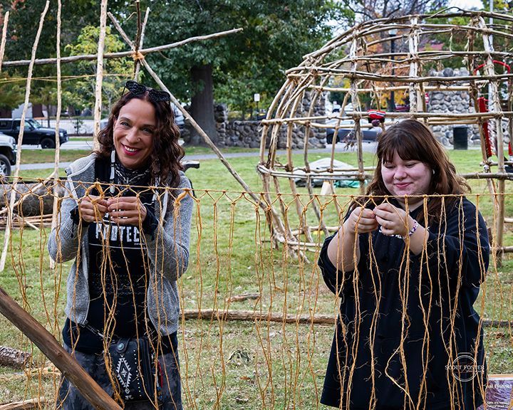 Two people weaving branches, constructing a wooden frame outdoors.