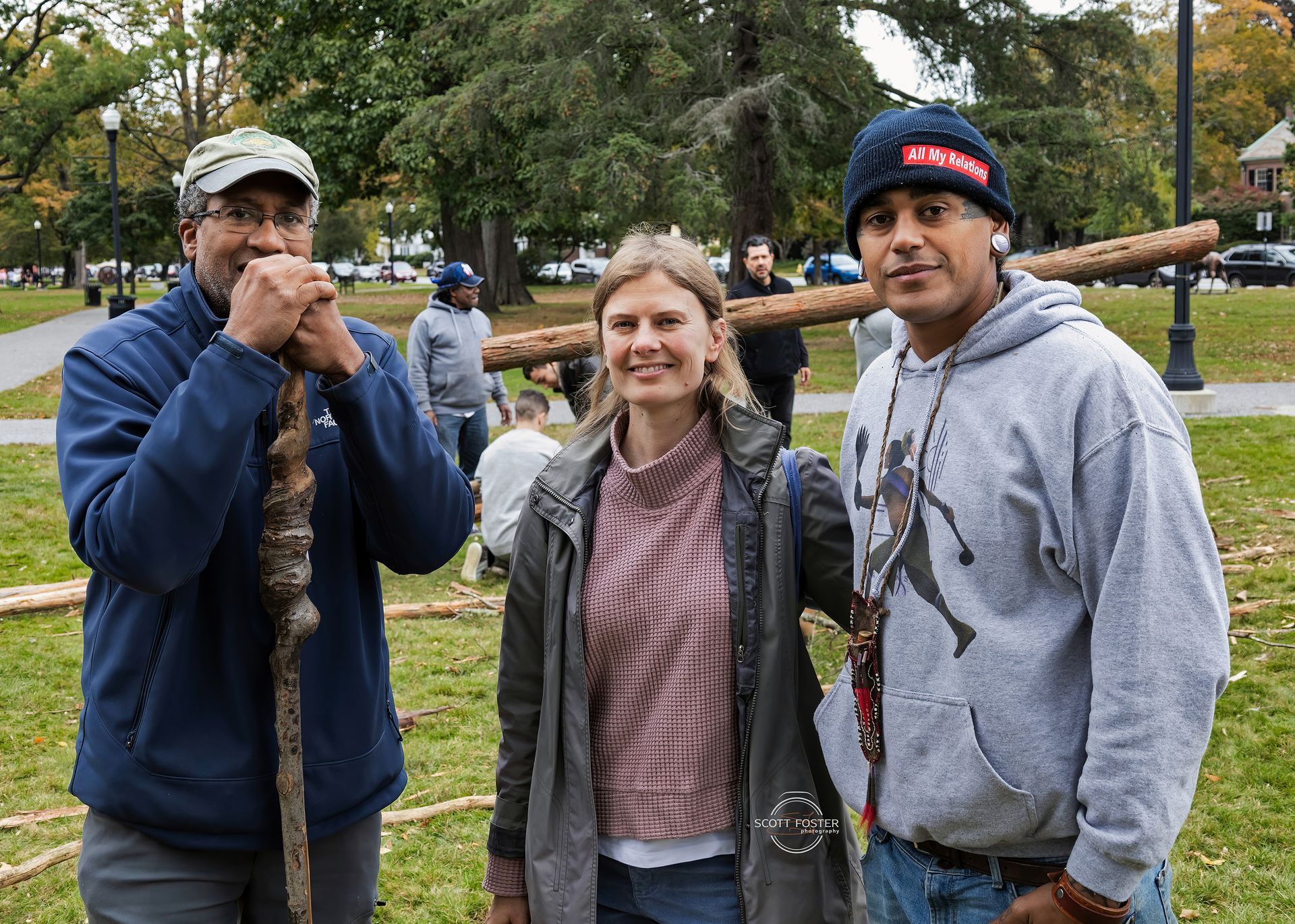 Three people in a park pose. One holds a staff, another stands between them, and a third carries a log.