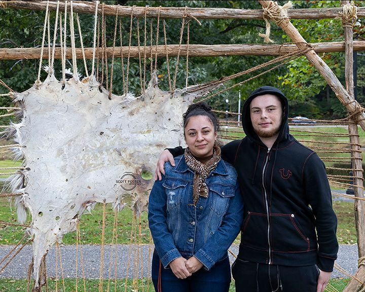Two people stand next to stretched animal hide drying on a wooden frame outdoors.
