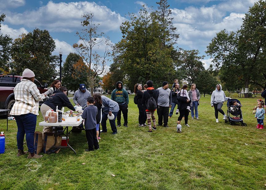 People gather around a table outdoors in a park. Some are looking at items on the table.