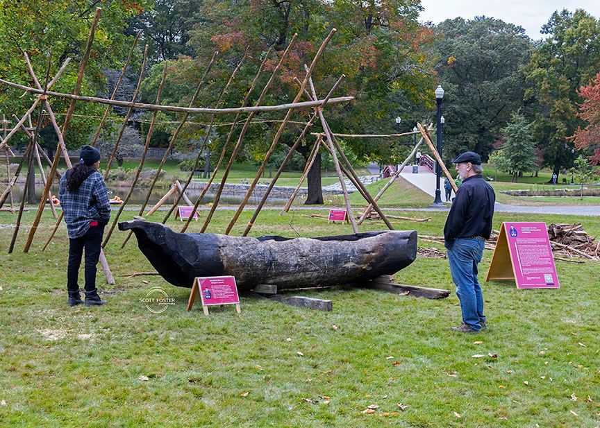 Two people looking at a large, charred wooden boat on grass, under a wooden frame, in a park.