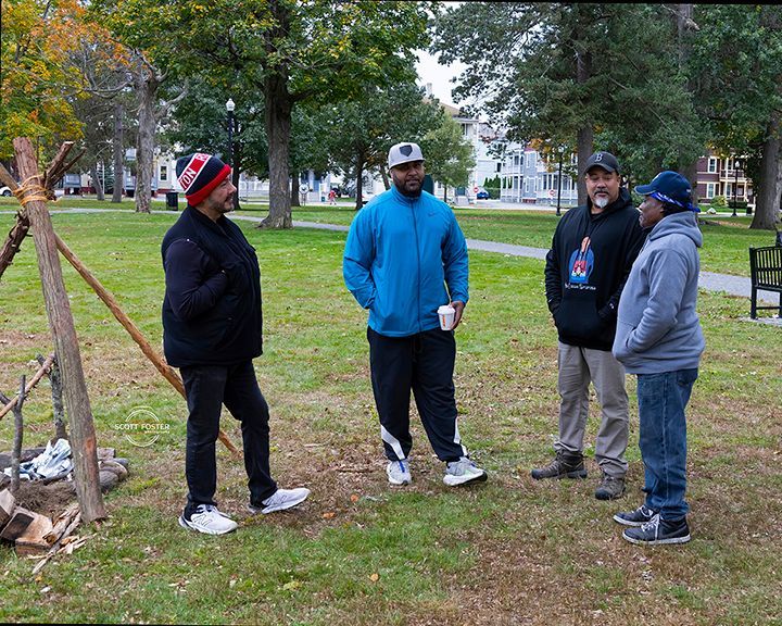 Four people stand in a park, conversing near a small teepee-like structure and fire pit.