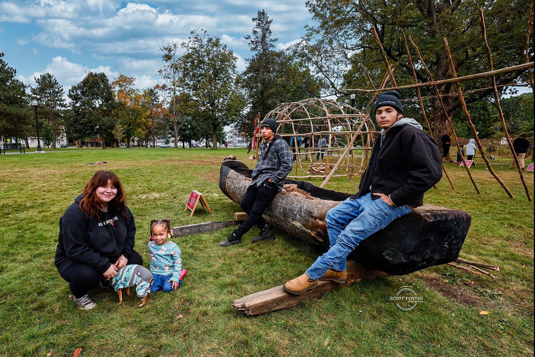 Family in park near wooden structures, sitting on log-like objects, with trees and cloudy sky background.