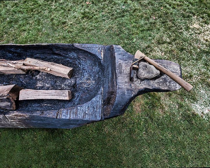 Charred wooden canoe, with logs inside, an axe and stone nearby, on green grass.