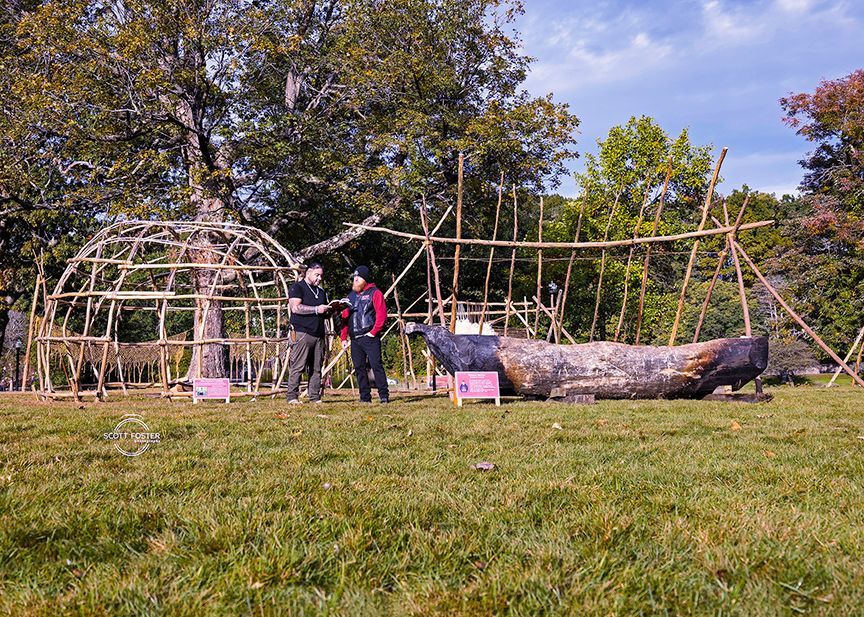 Two people stand near a wooden structure and a canoe on a grassy field.
