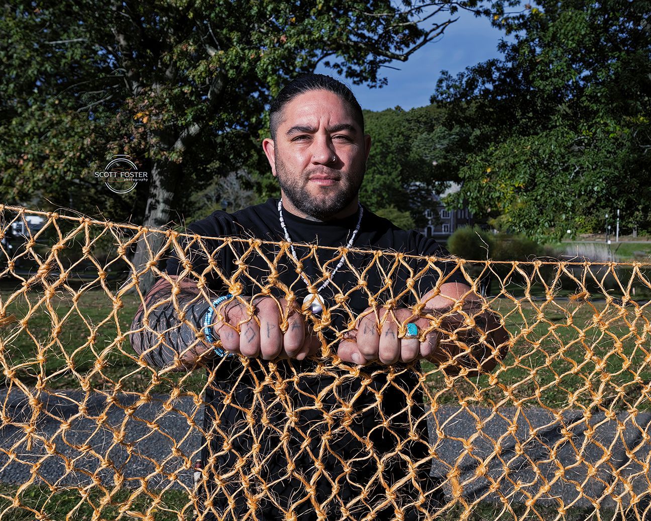 Man with tattoos behind a yellow net fence, looking at the viewer with trees and a park visible in the background.