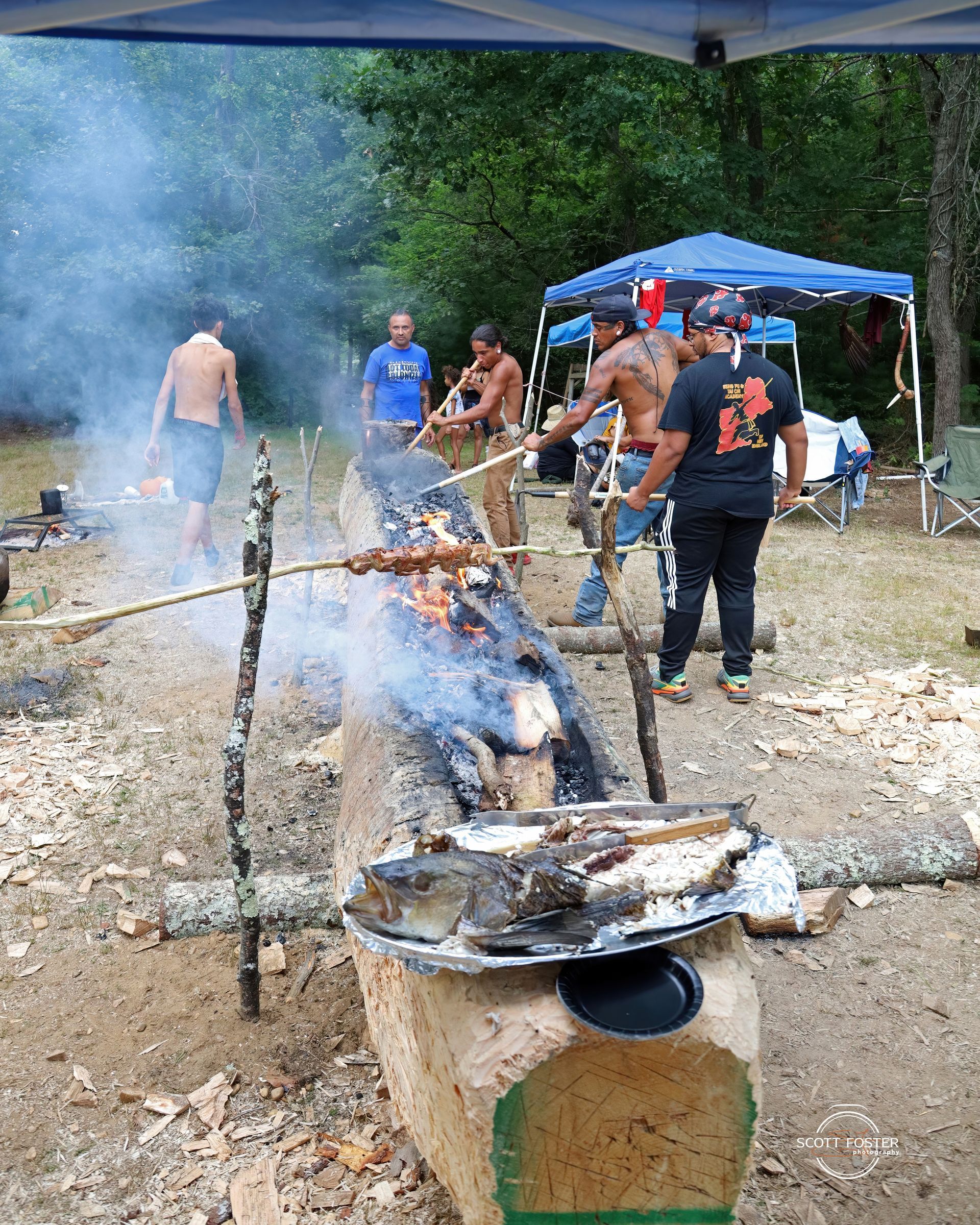 People cooking food over an open fire pit made from a hollowed log outdoors, smoke rising.