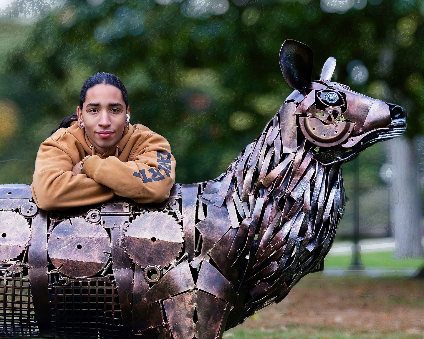 Man with arms crossed leans on a metal deer sculpture in an outdoor setting.