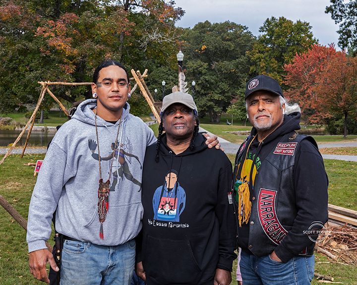 Three men pose outdoors, two with arms around each other. One wears a hoodie, one a vest with patches, and one a cap. 