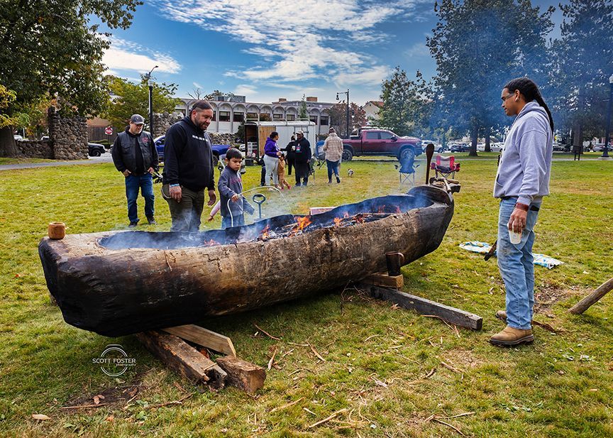 Large wooden canoe-like structure with fire inside, on grass. Several people watch.