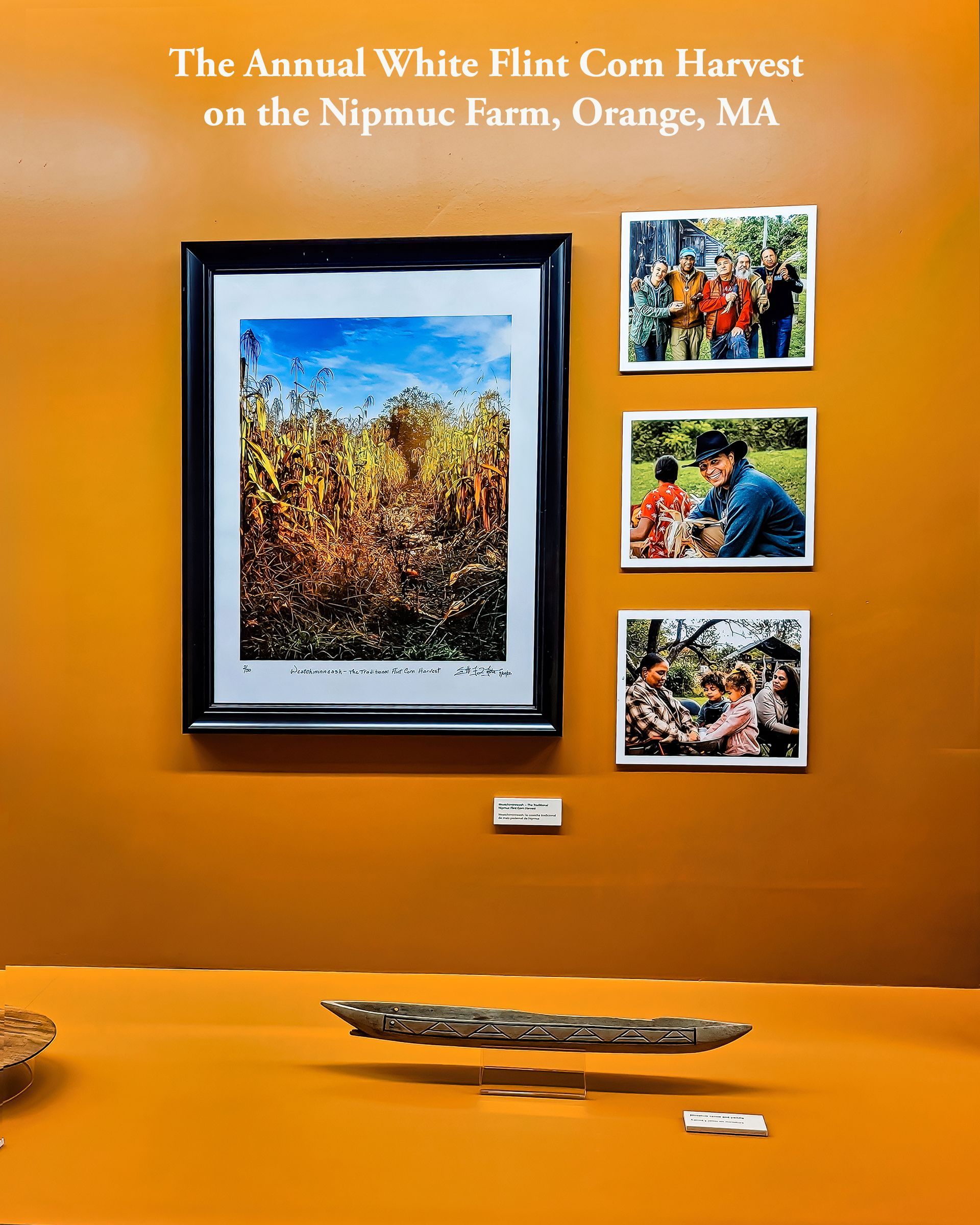 Display on an orange wall: framed cornfield art, three photos of people, and a wooden artifact.