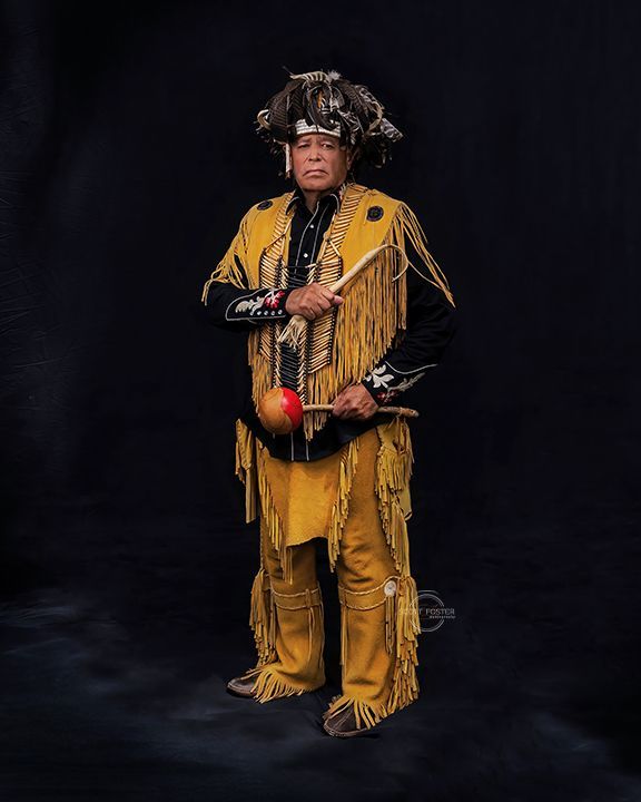 Man in yellow fringed outfit and headdress holds a drum, posing against black background.