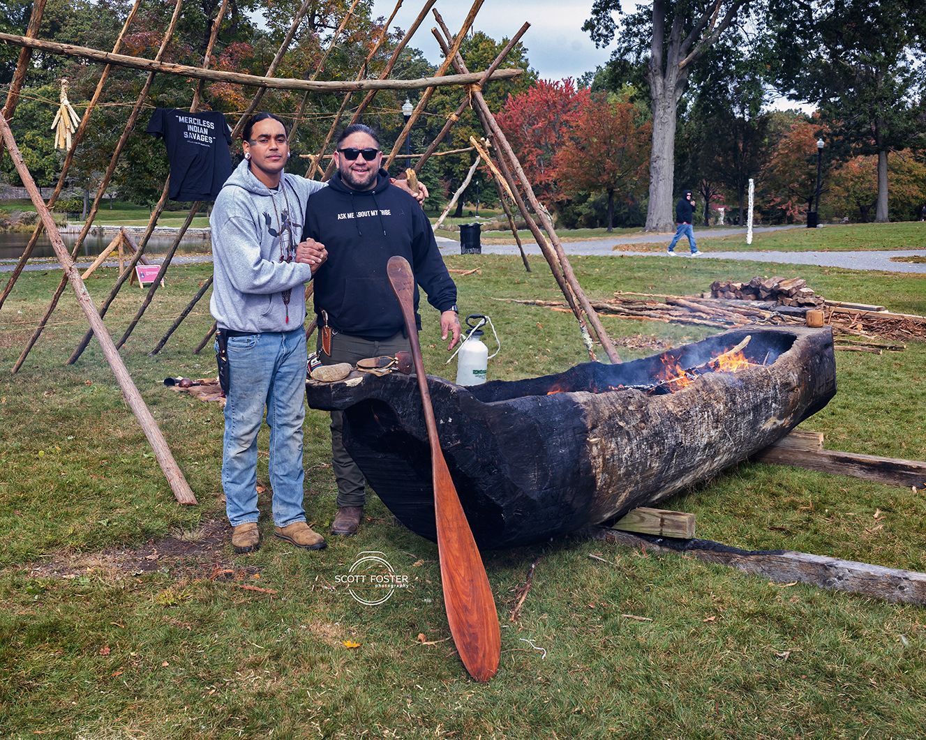 Two men stand beside a burning canoe, a wooden structure behind them. Outdoor setting, fall foliage.