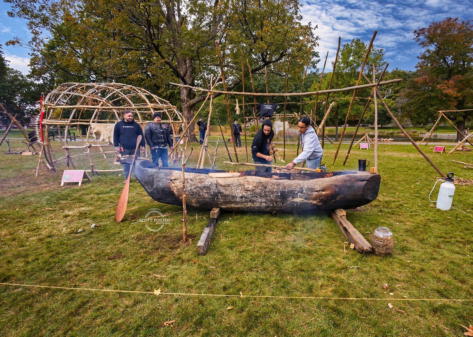 People around a carved wooden boat on grass, with structures of wooden sticks in the background.