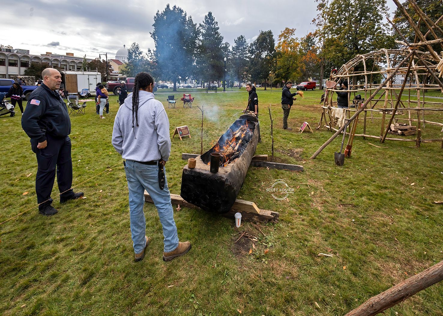 People watch a log burn on a grassy field; Native American structure, trees, and buildings are in the background.