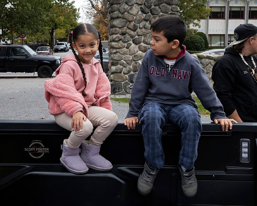 Two children sitting on a truck bed, one in pink smiling, one in blue looking to the side.