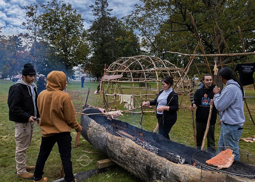 People gather around a large, charred log-canoe on grass, near a wooden structure.