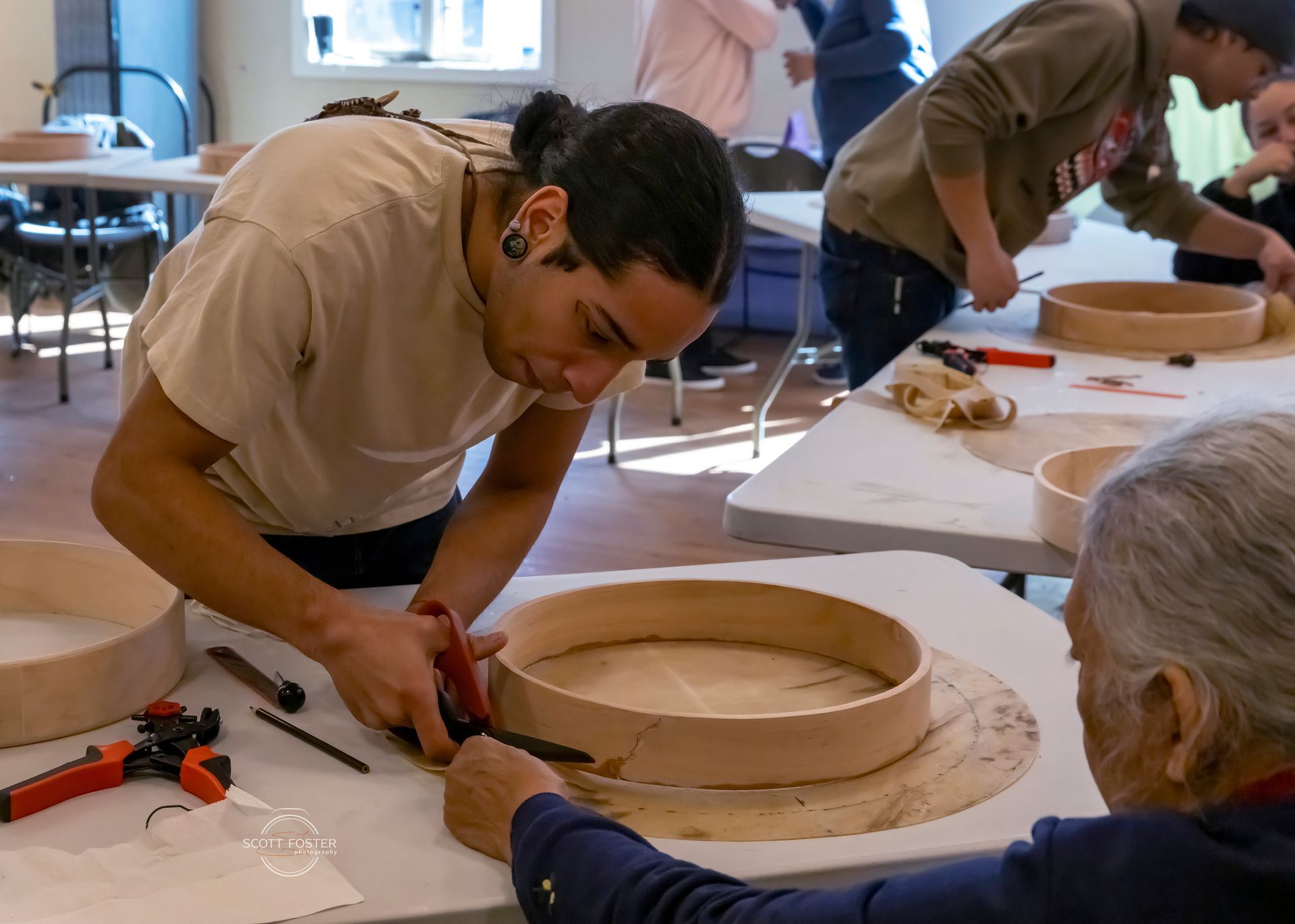 Man cutting leather, with others working on drum frames at a table. Indoor setting, bright.