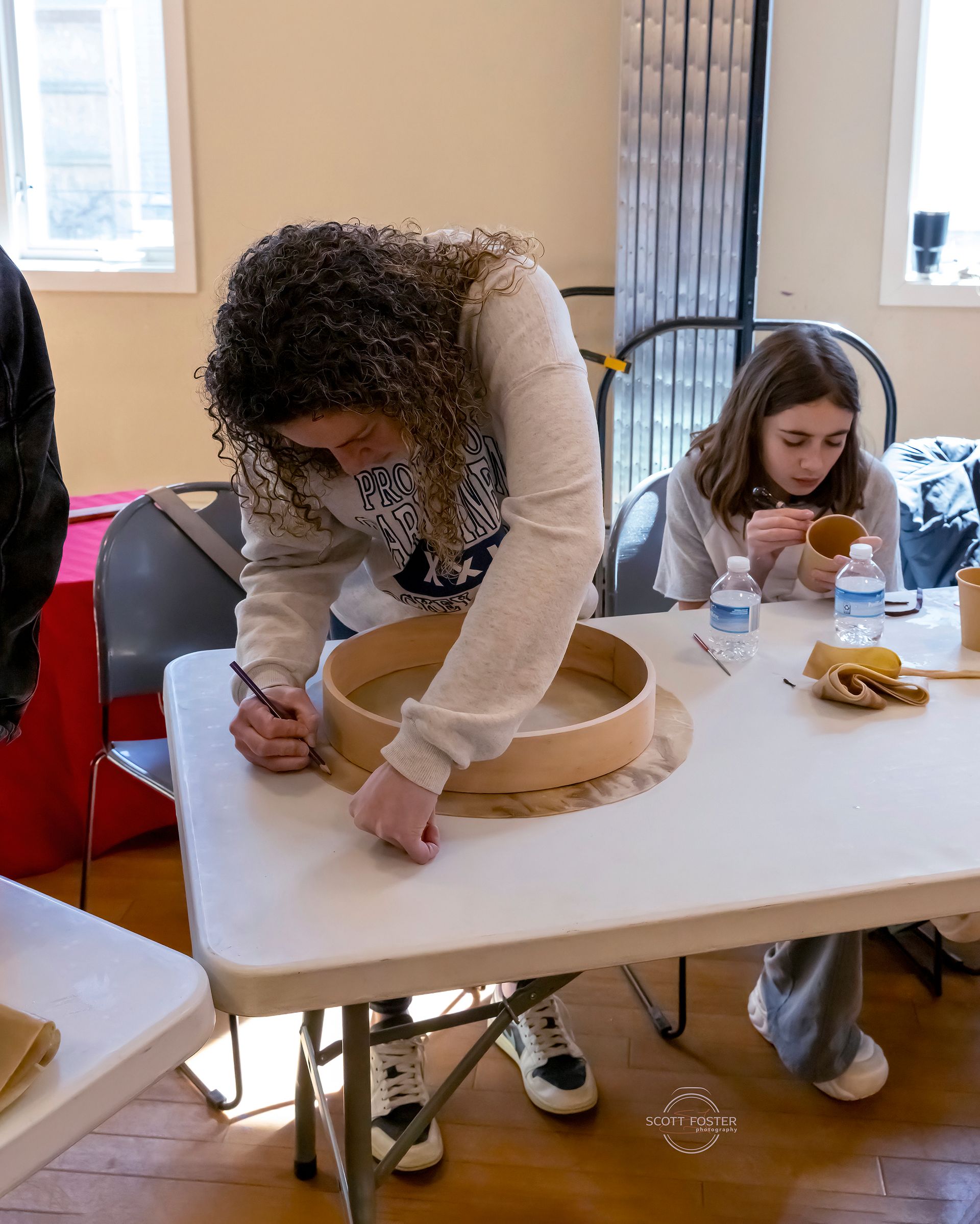 A person with curly hair draws on a wooden frame. Another person looks on. Indoors, crafting.