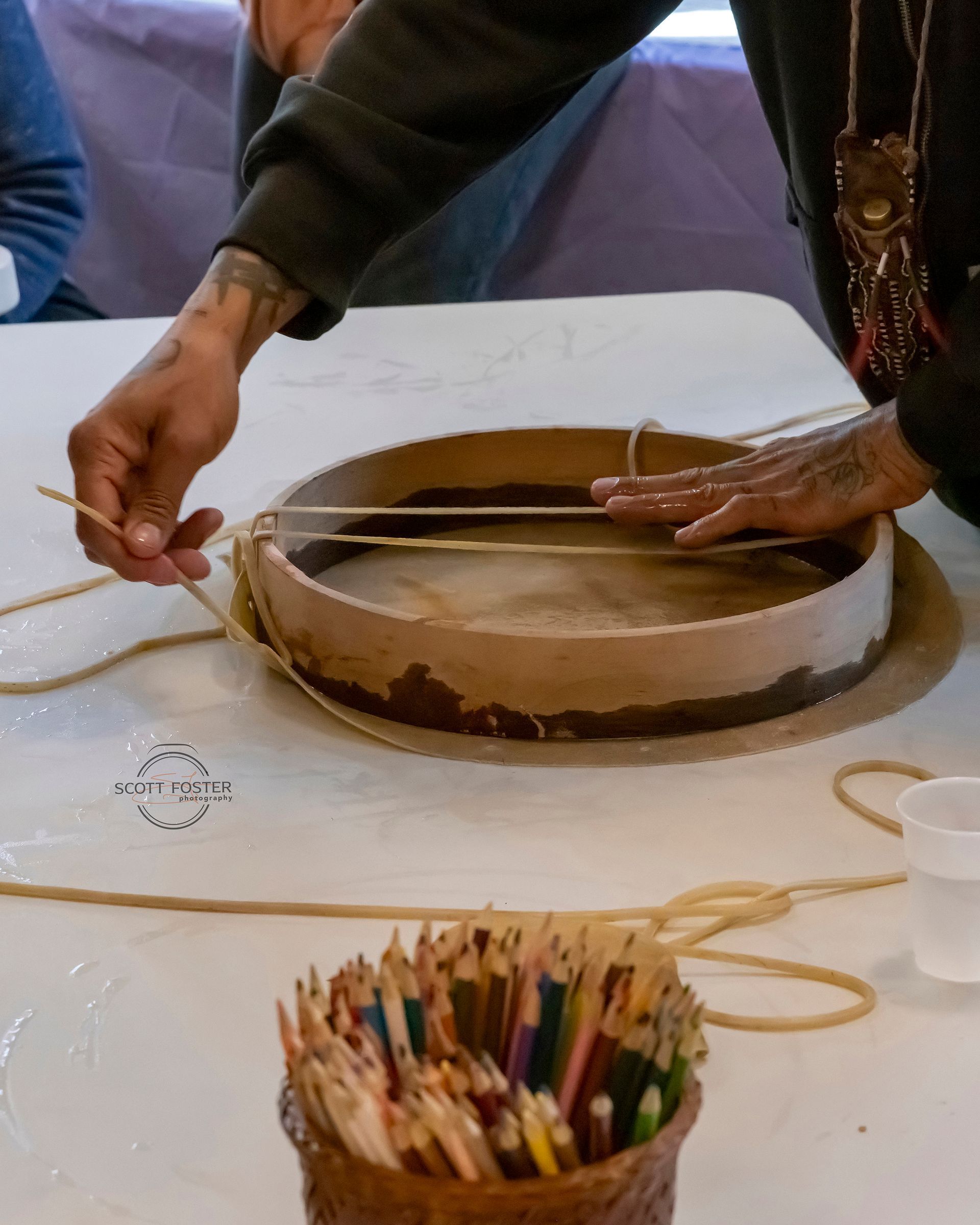 Person's hands working on a drum, alongside colored pencils and string on a white table.