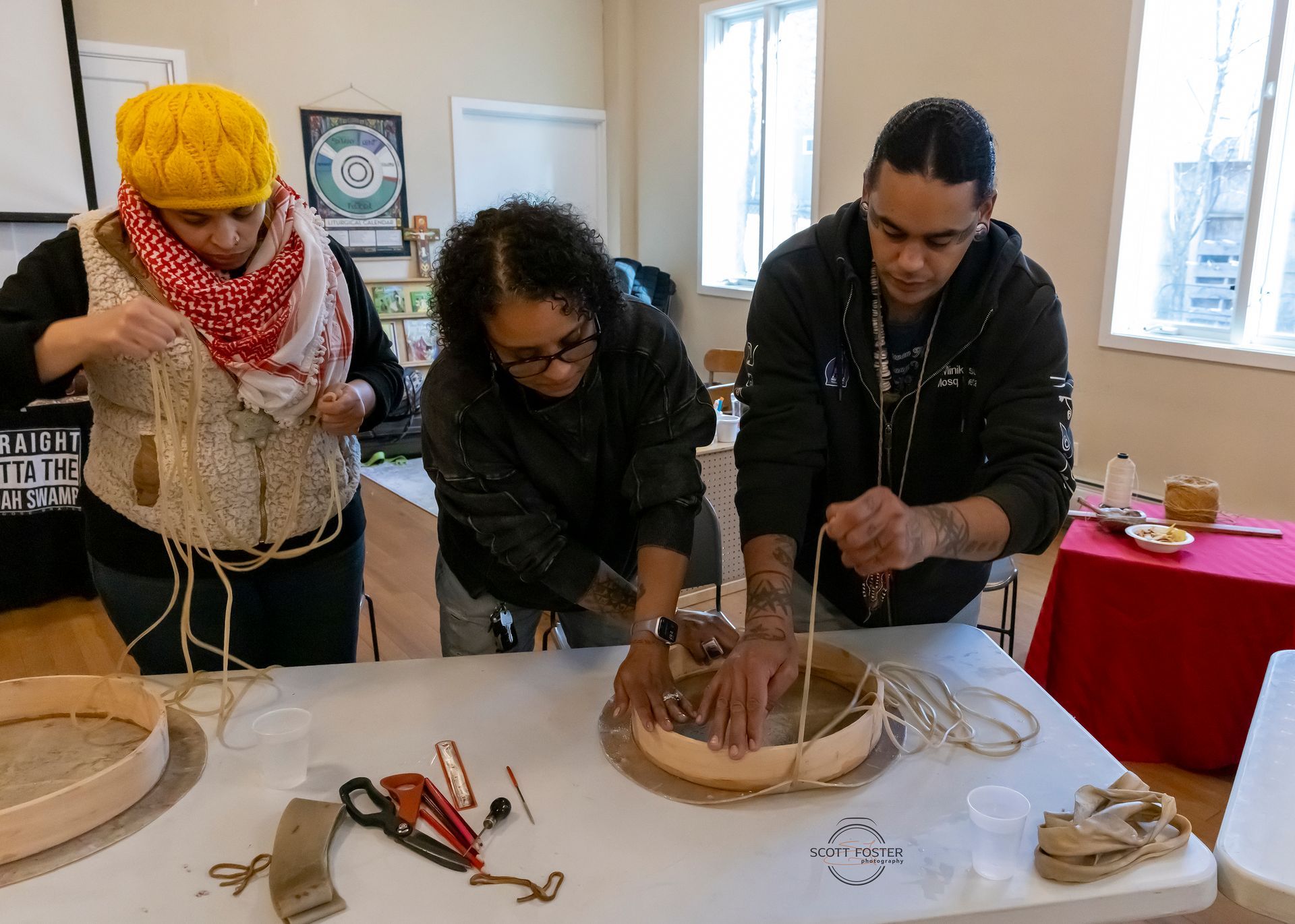 Three people crafting with string and a round wooden frame at a table.
