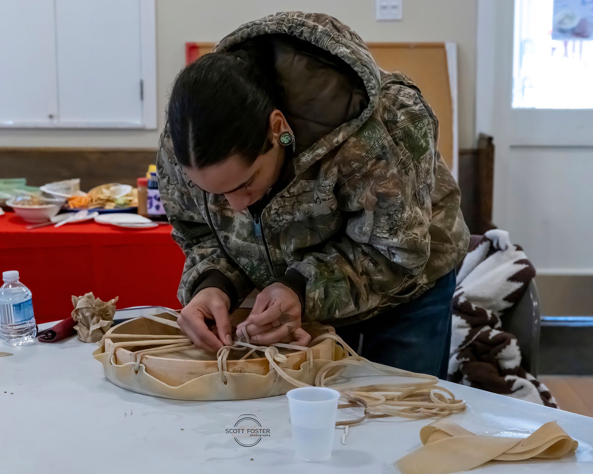 Person in camo jacket lacing a drum on a white table. Indoors.