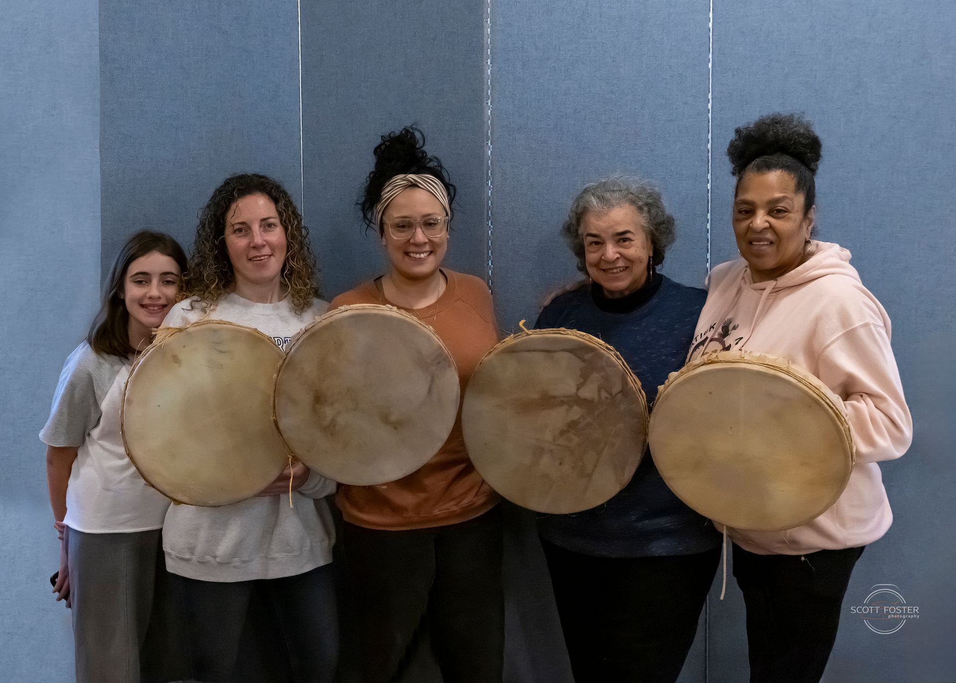 Five people holding drums, smiling, posing in front of a blue wall.