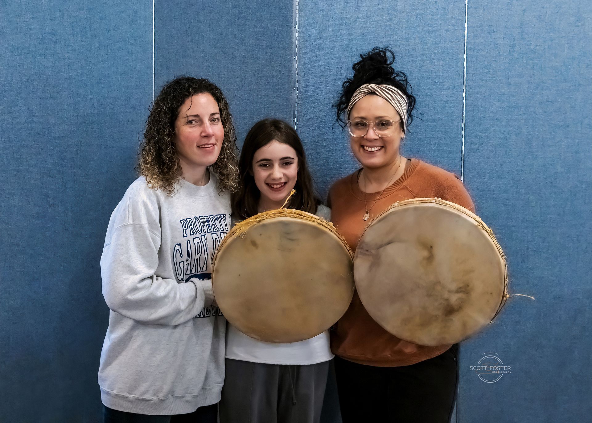 Three people holding drums in front of a blue wall. Two women and a child smile.