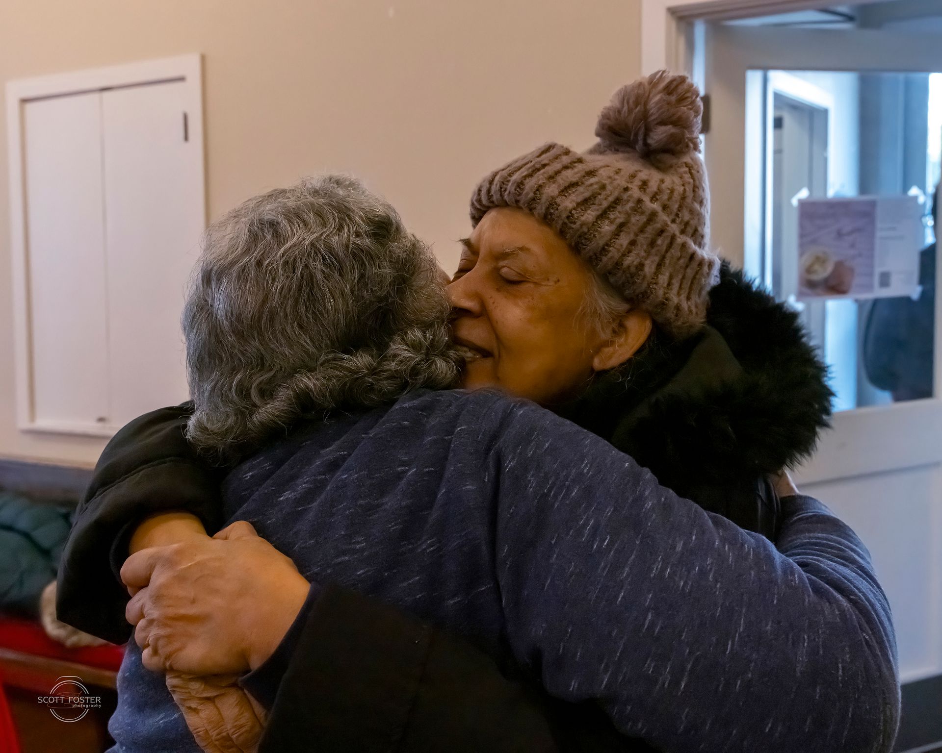 Two people embracing, one wearing a knit hat, in a well-lit indoor setting.