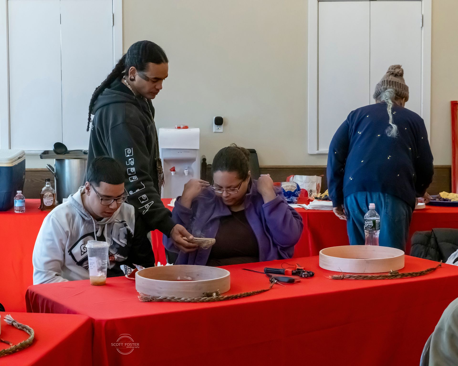 People gathered at a table crafting, beads and supplies present. One person is assisting another. 