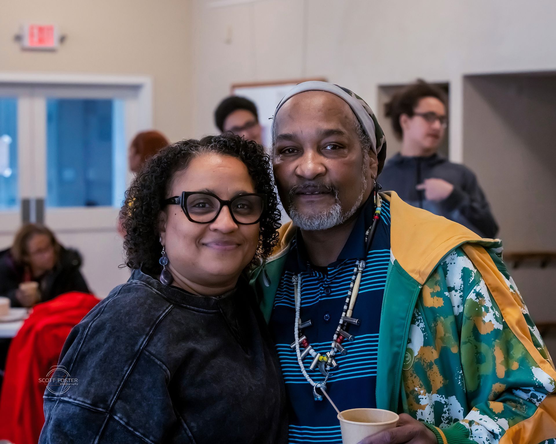 Two people smiling, posing together indoors. Woman with glasses, man with hat and colorful jacket. Others in background.