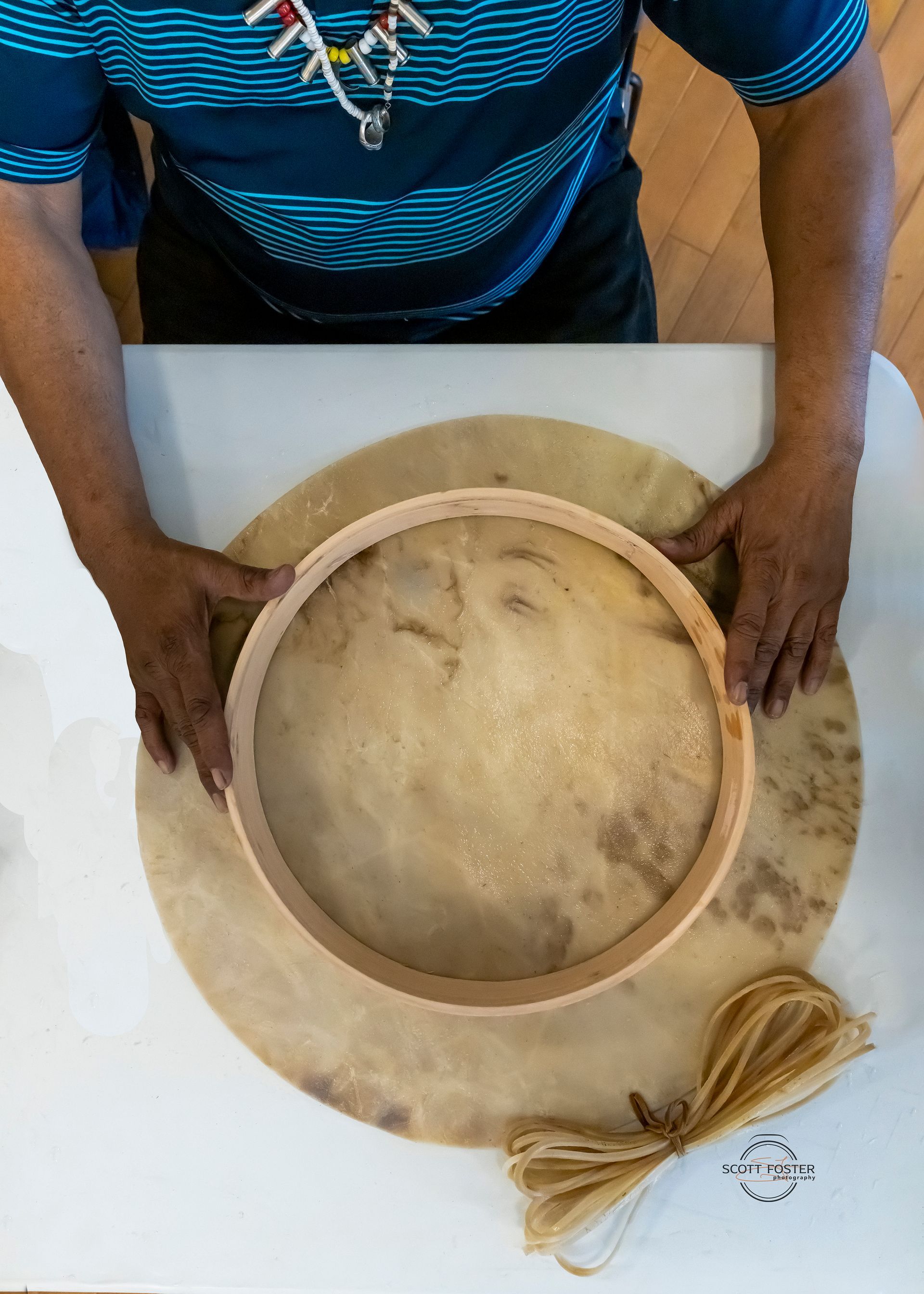 Person holding a drum frame over a large, round drum. Rawhide drum is light brown; frame is wood.
