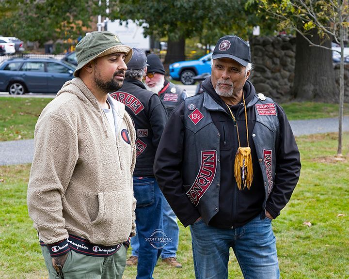 Two men wearing casual clothing are in a grassy outdoor area. One has a hat, the other a vest with patches.