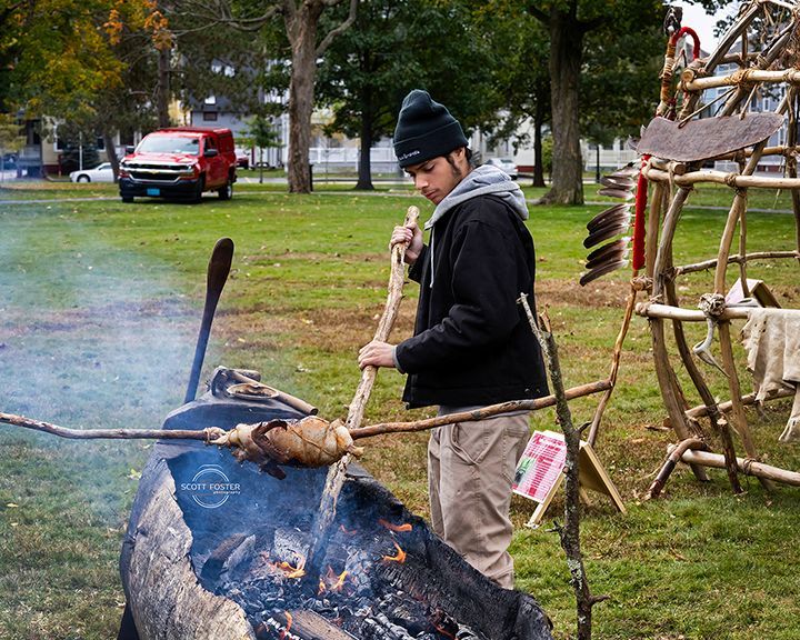 Man tending fire, roasting food on a spit in a park. Red van and wooden structure in background.