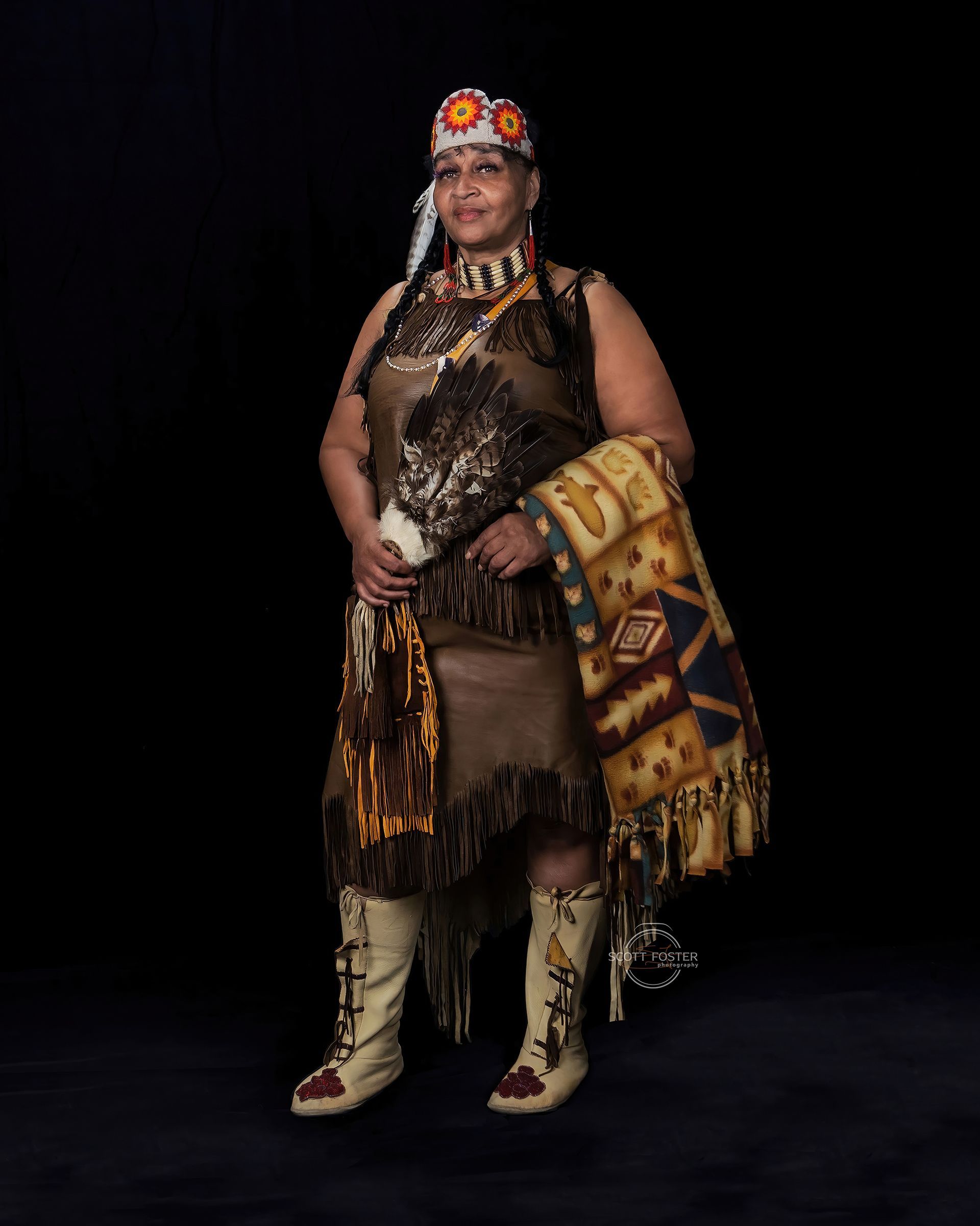 Woman in traditional Native American attire poses against a black background, holding a blanket.