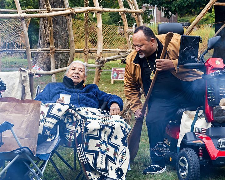 Two people near a small fire and wooden structure; one sits, wrapped in a blanket.