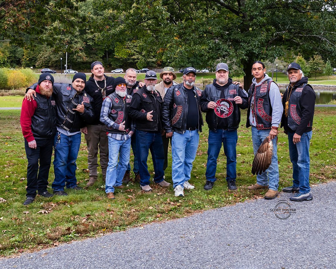 Group of men in vests and jeans pose outdoors. Some hold a motorcycle part and a bird.