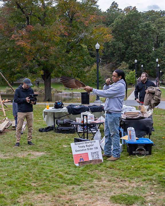 Man speaking at a park event, pointing. A camera operator films. Tables and signs are nearby.