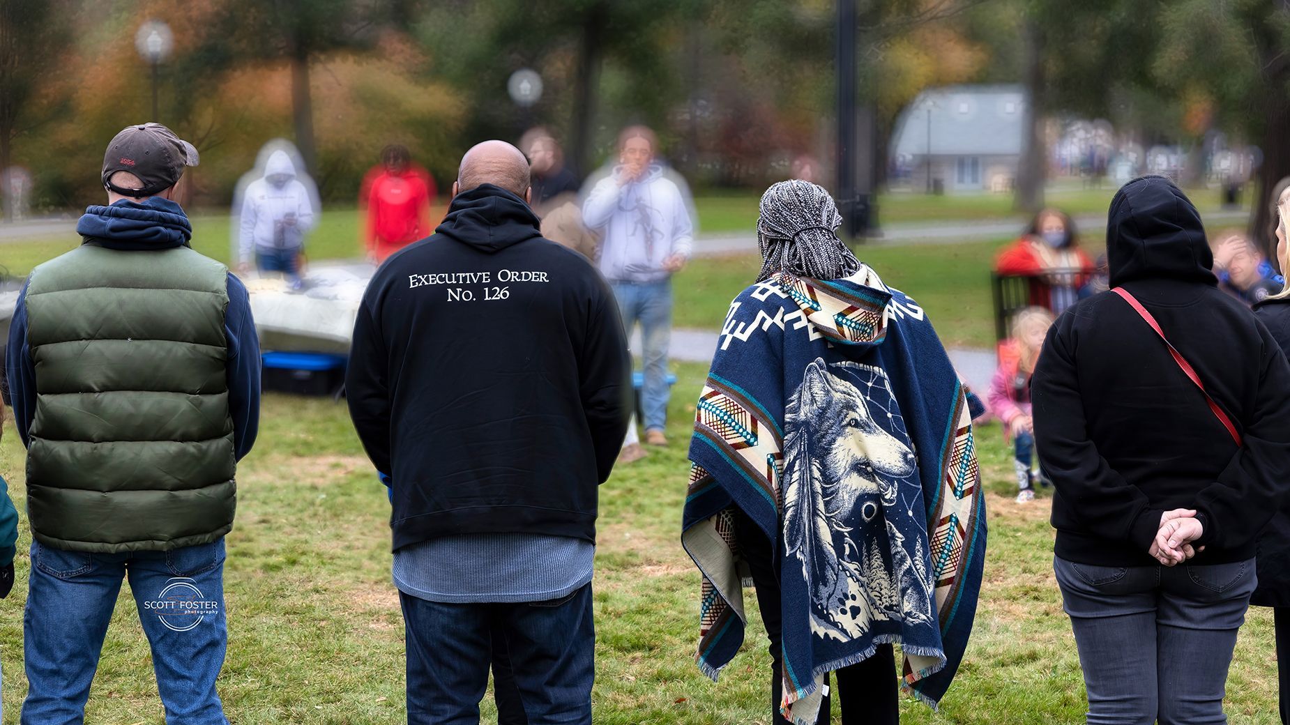 People standing in a park, facing away from the camera. Some wearing jackets or sweaters; fall foliage.