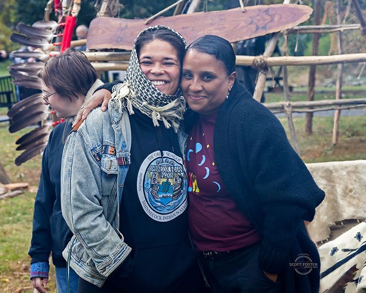 Two people smiling, arm in arm, pose outdoors. One wears a headscarf, denim jacket; the other a dark sweater.