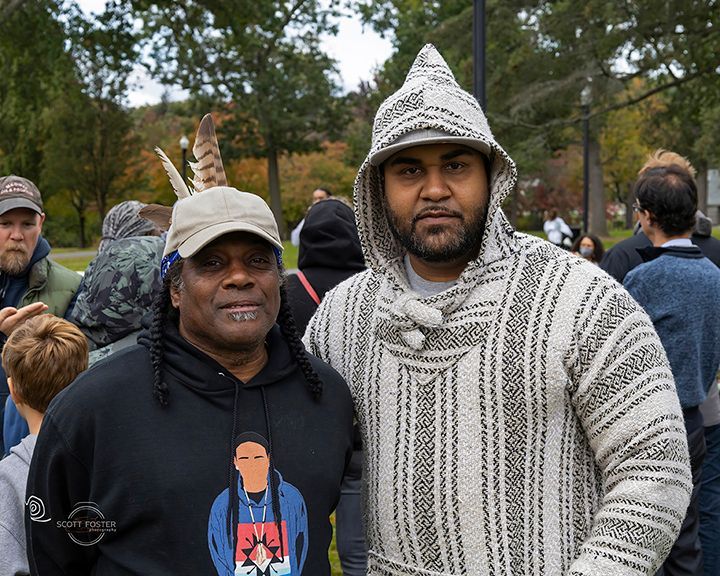 Two people pose outdoors. One wears a black sweatshirt with a graphic, the other a hooded sweater.