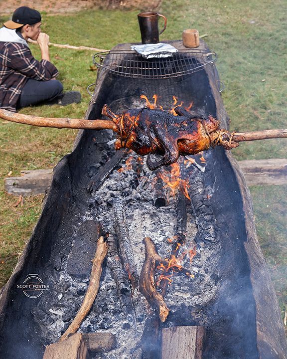 Chicken roasting over an open fire pit. Person sits nearby, observing.