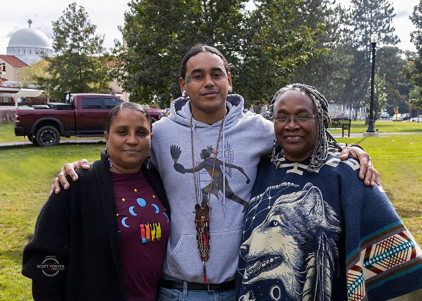 Three people pose outdoors, arms around each other. One wears a hoodie with a figure on it, others wear sweaters. 