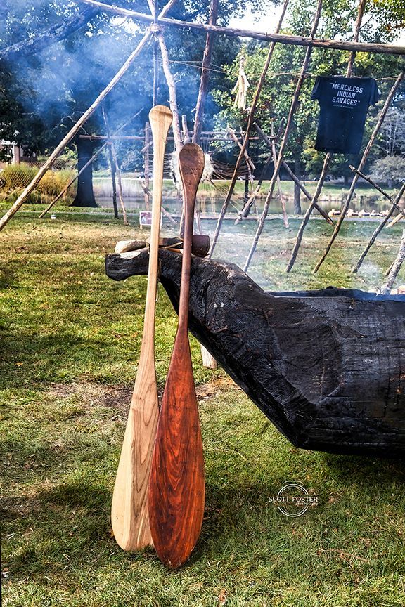 Two wooden canoe paddles hanging near a dark wooden canoe, outdoor setting.